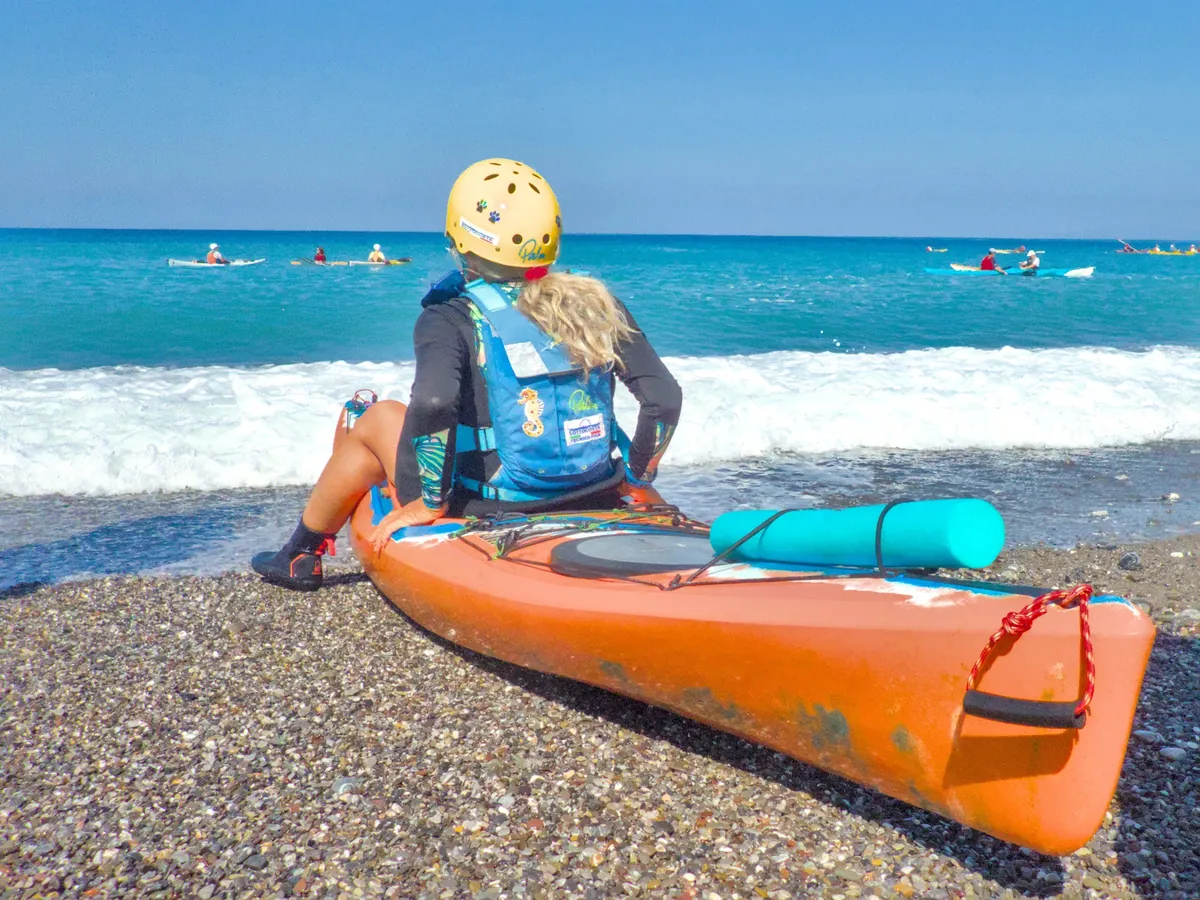 Attrezzatura kayak da mare: una pagaiatrice con casco e aiuto al galleggiamento seduta accanto al kayak, con la sacca stagna in coperta