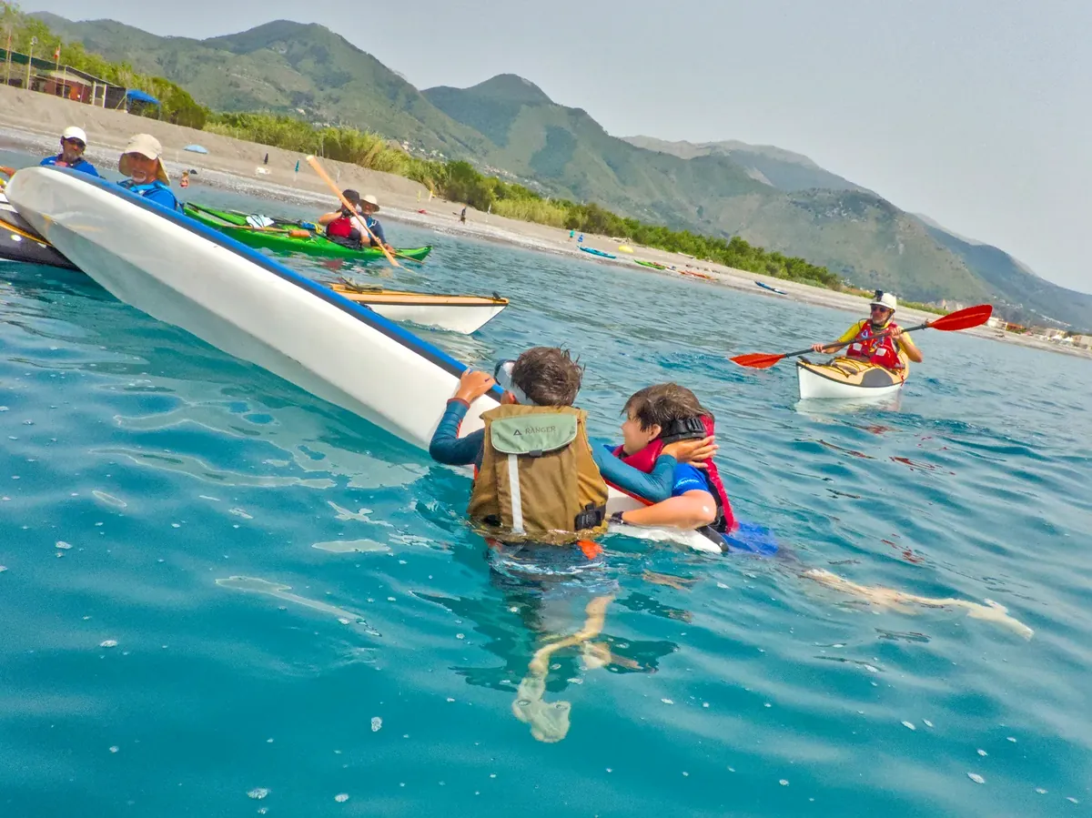 Corso di kayak da mare: due ragazzi in acqua accanto al kayak rovesciato durante un’esercitazione di autosoccorso, con altri pagaiatori vicino in kayak