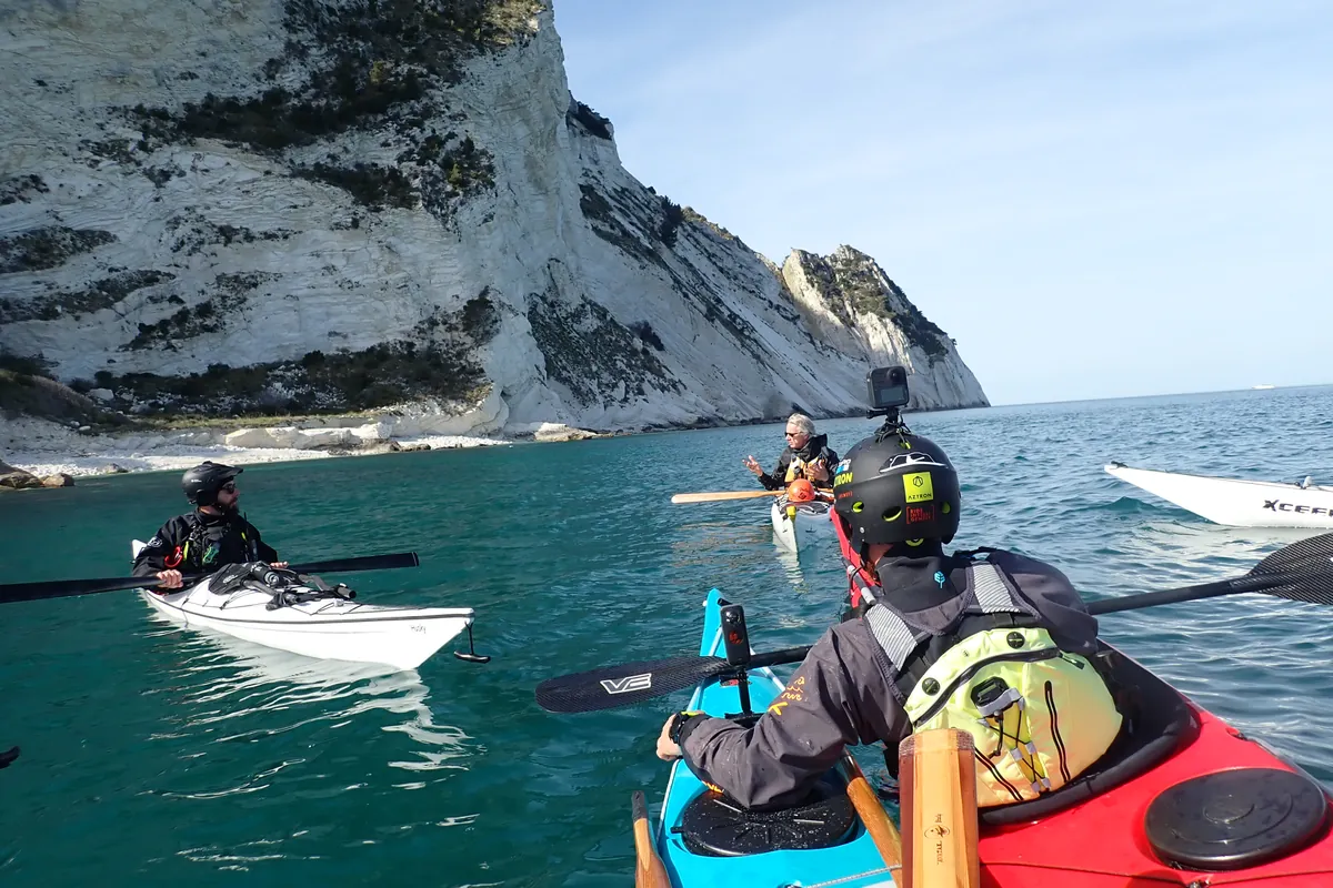 Esame Pagaia Azzurra: pagaiatore con casco e videocamera che segue il gruppo sotto una falesia bianca