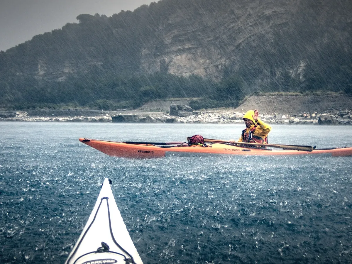 Meteo mare per il kayak: pagaiatore con giacca impermeabile sotto la pioggia battente, vicino alla costa